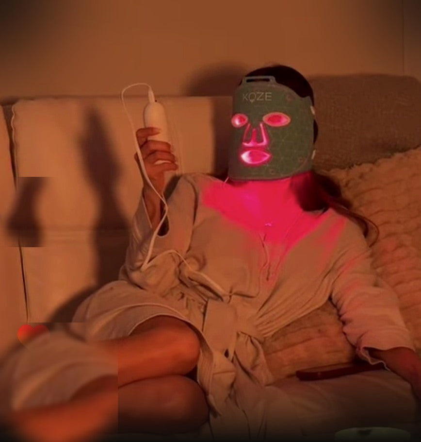 A person in a robe on a cream sofa at night, wearing a gray KOZE LED face mask glowing bright red, holding the remote.
