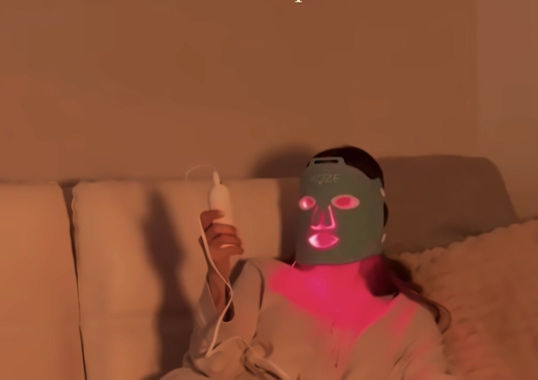 A person in a robe on a cream sofa at night, wearing a gray KOZE LED face mask glowing bright red, holding the remote.